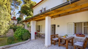 A covered patio with a stone floor, wooden furniture, and a view of a white building with shutters. - Amazing apartment in Mundanije (Mundanije)