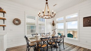 A dining area with a wooden table, six chairs, a chandelier, and large windows with blinds.