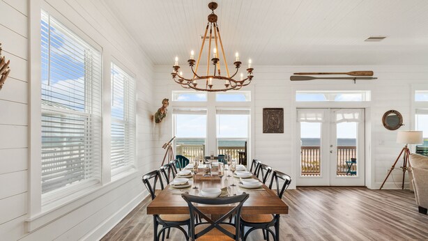 A dining room with a wooden table set for six, surrounded by chairs, a chandelier, and a view of the ocean through large windows.