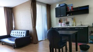 Una sala de estar con un sofá, una mesa de comedor con sillas, una zona de cocina con armarios y un fregadero, y una ventana con cortinas.