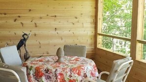 Eine Holzdachterrasse mit einem runden Tisch und vier Stühlen, einem Deckenventilator und einem Fenster mit Blick auf Bäume.