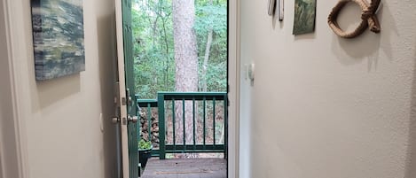 Un pasillo con piso de madera, barandal verde y vista a un bosque a través de una puerta abierta.