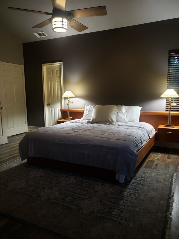 A bedroom with a bed, two bedside lamps, a ceiling fan, a door, and a window with blinds.