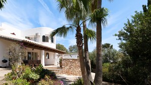 Una villa in stile mediterraneo con facciata in pietra, un patio con mobili da esterno e un rigoglioso giardino con palme.