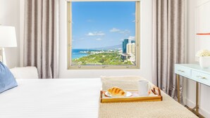 A hotel room with a bed, a bedside table with a lamp, a window with a view of a cityscape and the sea, and a tray with a croissant and a cup. - Ocean-City Views Glamorous Condo at The Watermark w/Parking & Full Kitchen (Honolulu)