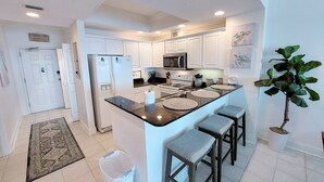 A modern kitchen with white cabinets, a central island with stools, a refrigerator, stove, and microwave. - Crystal Shores1006 2 Bedroom Condo by RedAwning (Gulf Shores)