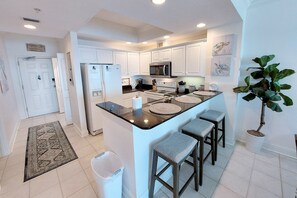 A modern kitchen with white cabinets, a central island with stools, a refrigerator, stove, and microwave. - Crystal Shores1006 2 Bedroom Condo by RedAwning (Gulf Shores)