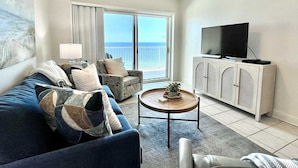 A living room with a sofa, armchairs, a coffee table, a TV, and a view of the sea through large windows. - Crystal Shores1006 2 Bedroom Condo by RedAwning (Gulf Shores)
