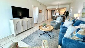 A living room with a TV, sofa, and coffee table. There is a kitchen in the background. - Crystal Shores1006 2 Bedroom Condo by RedAwning (Gulf Shores)