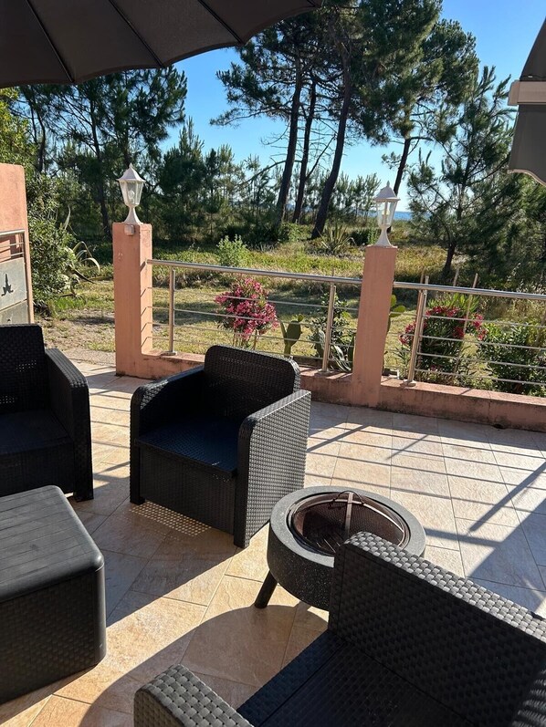 Une terrasse extérieure aménagée avec des meubles en osier, un foyer et une vue sur des arbres et un bâtiment en arrière-plan.