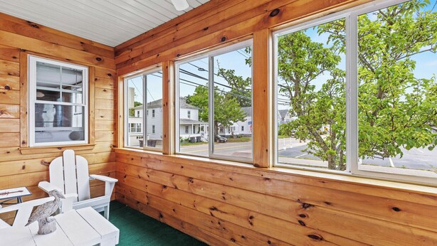 A wooden cabin with a covered porch, white Adirondack chairs, and a view of a street and trees.