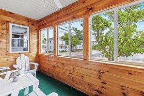 Una cabaña de madera con porche cubierto, sillas Adirondack blancas y vista a la calle y árboles.