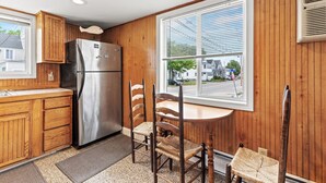 A kitchen with wooden cabinets, a stainless steel refrigerator, a dining table with chairs, and a window with blinds.