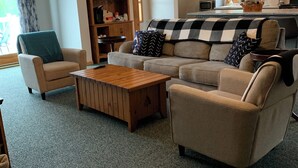 Ein gemütliches Wohnzimmer mit einer Couch, einem Sessel und einem Couchtisch. Ein Bücherregal mit verschiedenen Gegenständen und ein Fenster mit Vorhängen sind ebenfalls vorhanden.