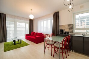 A modern apartment with a red sofa, a glass dining table with red chairs, a kitchen with a black countertop, and large windows offering a city view.