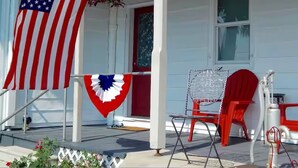 A porch with an American flag, a red chair, and a clear plastic chair.