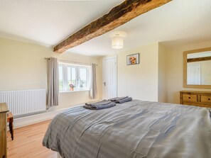 A bedroom with a bed, a window with curtains, a radiator, a wooden dresser, and a ceiling beam.