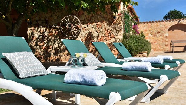 A row of green lounge chairs with towels and a tray with glasses and lemons, set against a stone wall with a wooden wheel and greenery.