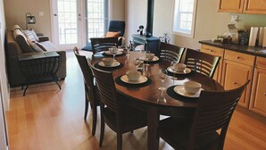 A dining area with a round table set for six, surrounded by wooden chairs. A kitchen with wooden cabinets and a stove is visible in the background. - The Cabin on Grey Owl w/hot tub (Onanole)