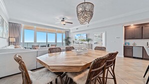 A spacious living room with a large wooden dining table, wicker chairs, a ceiling fan, and a view of the ocean through large windows. - New Complex!  Lazy River/Slide, Beachfront! (Orange Beach)