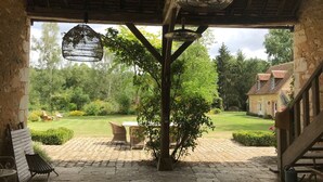 A covered outdoor area with a stone floor, wooden furniture, and a view of a garden and house in the background. - 14-person family home in Perche with heated pool and tennis court (Rémalard-en-Perche)