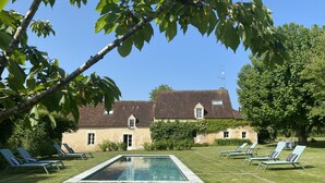 A pool area with lounge chairs, a stone building with a tiled roof, and a clear blue sky. - 14-person family home in Perche with heated pool and tennis court (Rémalard-en-Perche)