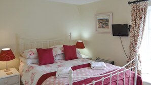 Un lit bien fait, avec un cadre en métal blanc, une literie rouge et une couverture rose à pois. On y voit une table de chevet blanche avec une lampe et un petit plateau en bois. Un téléviseur est fixé au mur, et une fenêtre avec un rideau à motifs floraux est visible.