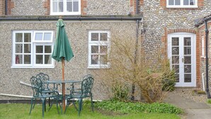 Un bâtiment en pierre avec deux fenêtres et une porte, un parapluie vert et une table avec des chaises au premier plan.