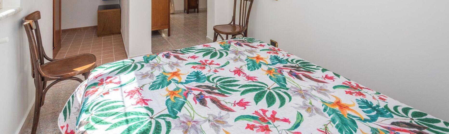 Quarto com uma cama, duas cadeiras de madeira, uma mesa pequena e uma porta que dá para outro cômodo.