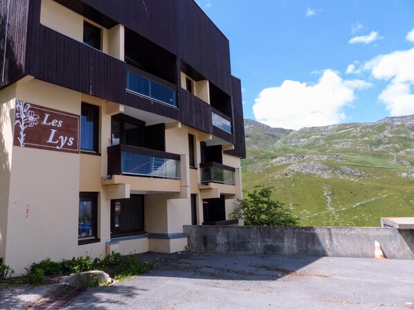 A multi-story building with balconies, a sign reading 'Les Lys,' and a mountainous landscape in the background.
