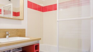 Een badkamer met een witte wastafel, een houten opbergmeubel en een rode plank, en een douche met een deur van geëtst glas.