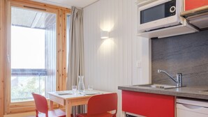 A compact kitchen with a microwave, sink, and dishwasher. There is a dining table with two chairs and a window with curtains. - travelski home select - Résidence Les Lys - Studio 2 personnes Bâtiment B (Les Menuires)