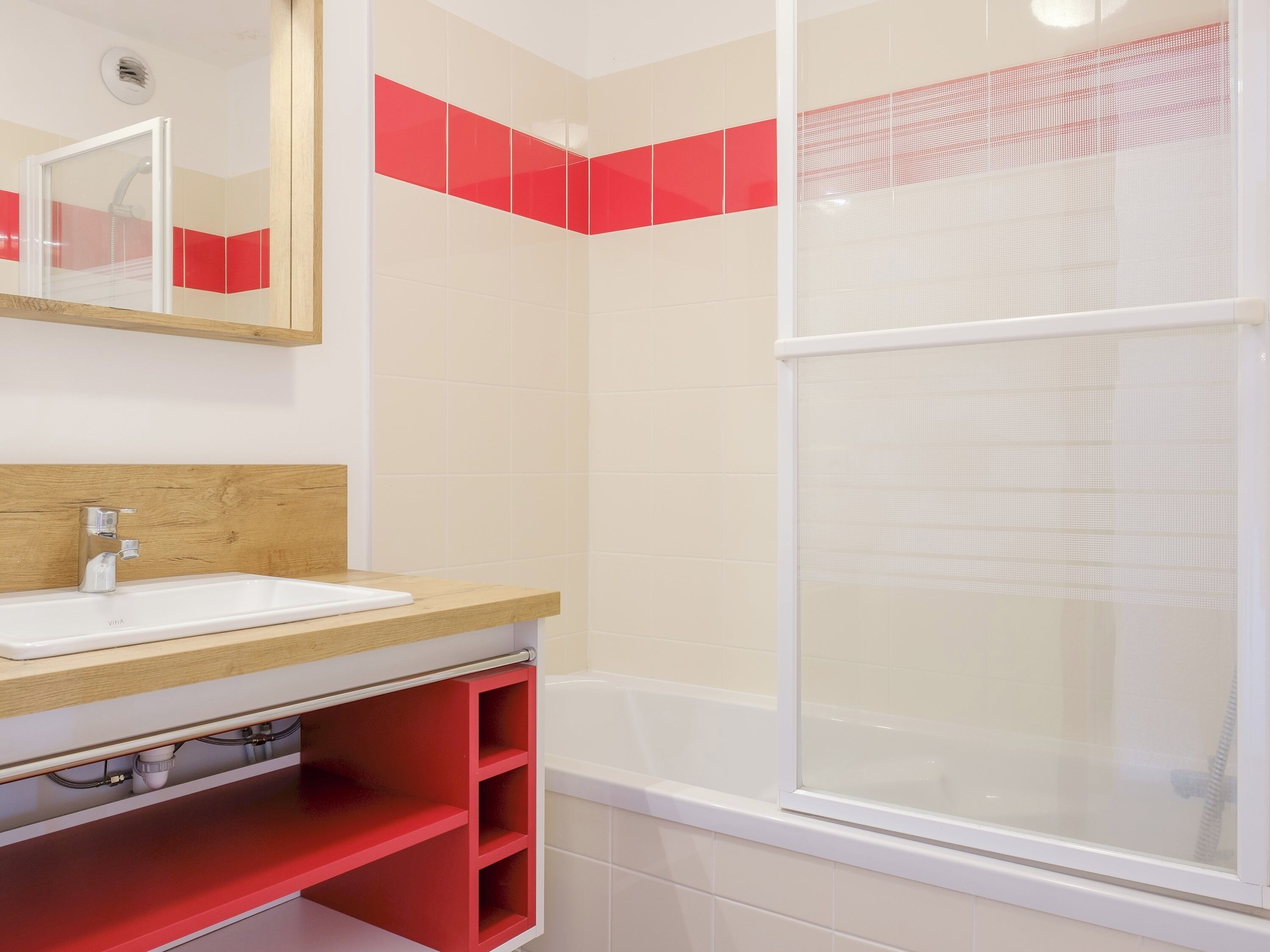 Een badkamer met een witte wastafel, een houten opbergmeubel en een rode plank, en een douche met een deur van geëtst glas.