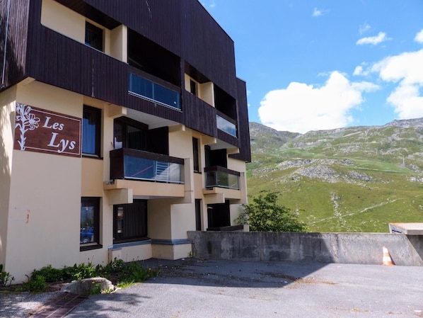 A multi-story building with balconies, a sign reading 'Les Lys,' and a mountainous landscape in the background.