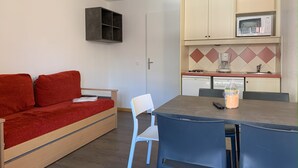 Ein kompakter Wohnraum mit einem roten Sofa, einem kleinen Esstisch mit Stühlen, einer Küchenzeile mit Mikrowelle und Geschirrspüler.