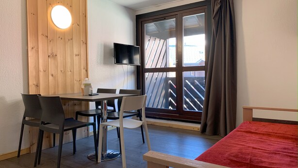 Ein Zimmer mit Holzwand, einem Fernseher über einer Schiebetür aus Glas, einem Bett mit roter Bettdecke und einem Tisch mit Stühlen.