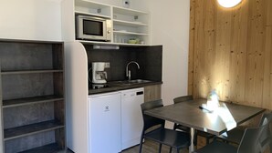 Cocina compacta con microondas, cafetera, pileta y heladera pequeña. Hay una mesa de comedor con sillas y un revestimiento de madera con una lámpara circular.