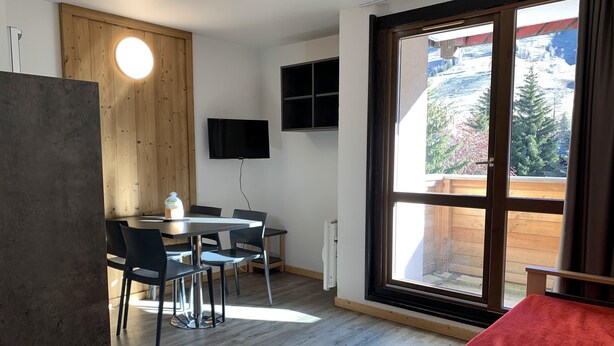 Una habitación moderna con piso de madera, una mesa de comedor con sillas, un televisor montado en la pared y un amplio ventanal con vista a montañas.