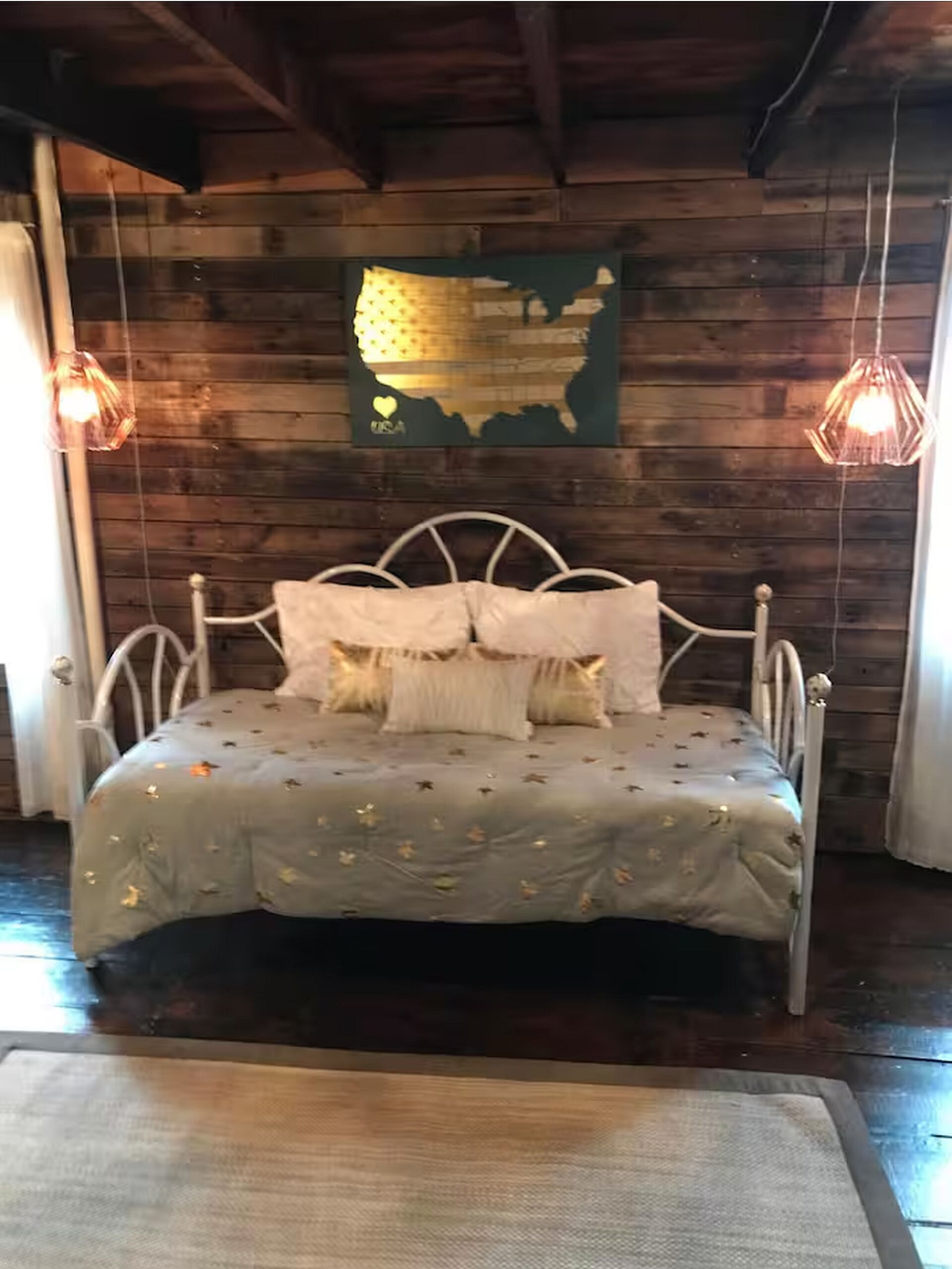 Una cama con estructura metálica y colcha gris con diseños de hojas, frente a una pared de madera con un mapa de los Estados Unidos.