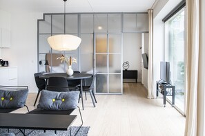 A modern living room with a dining area, a large window with curtains, and a television on a stand. - The Beach Gardens - Daniel&Jacob's Premium Apartments (Copenhagen)