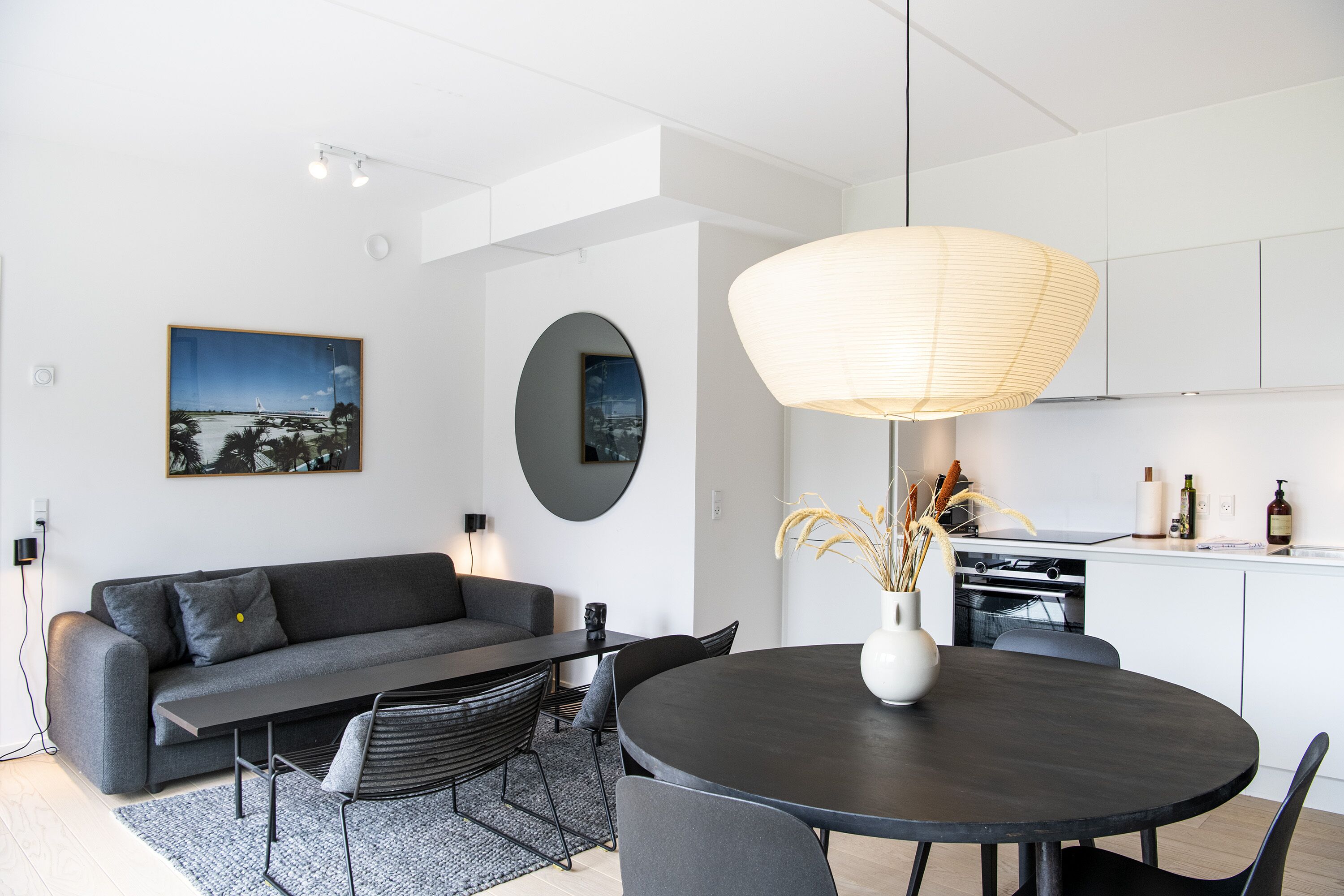A modern living room with a sofa, dining table, and chairs. There is a kitchen area with a stove and cabinets.
