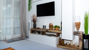 Ein modernes Wohnzimmer mit einem Flachbildfernseher an einer weißen Wand, einem Holztischchen mit Dekorationen, einem großen Fenster mit transparenten Vorhängen und einer kleinen Zimmerpflanze.