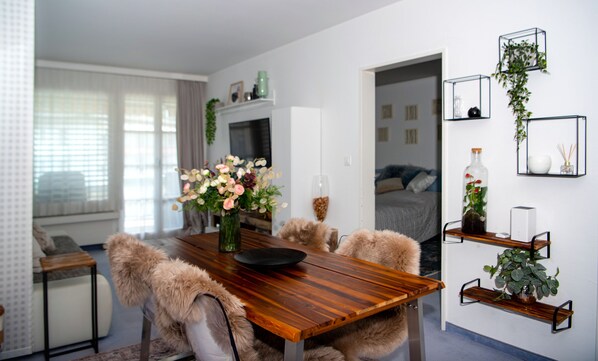 A modern living room with a wooden dining table, fluffy chairs, a TV, and a cozy bed in the background.