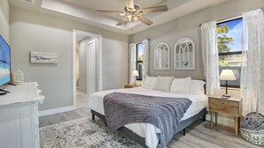A bedroom with a bed, nightstand, and a ceiling fan. There is a window with curtains, a wall-mounted TV, and a decorative sign. - Amazing Outdoor kitchen & Private Screened In Pool (Bonita Springs)