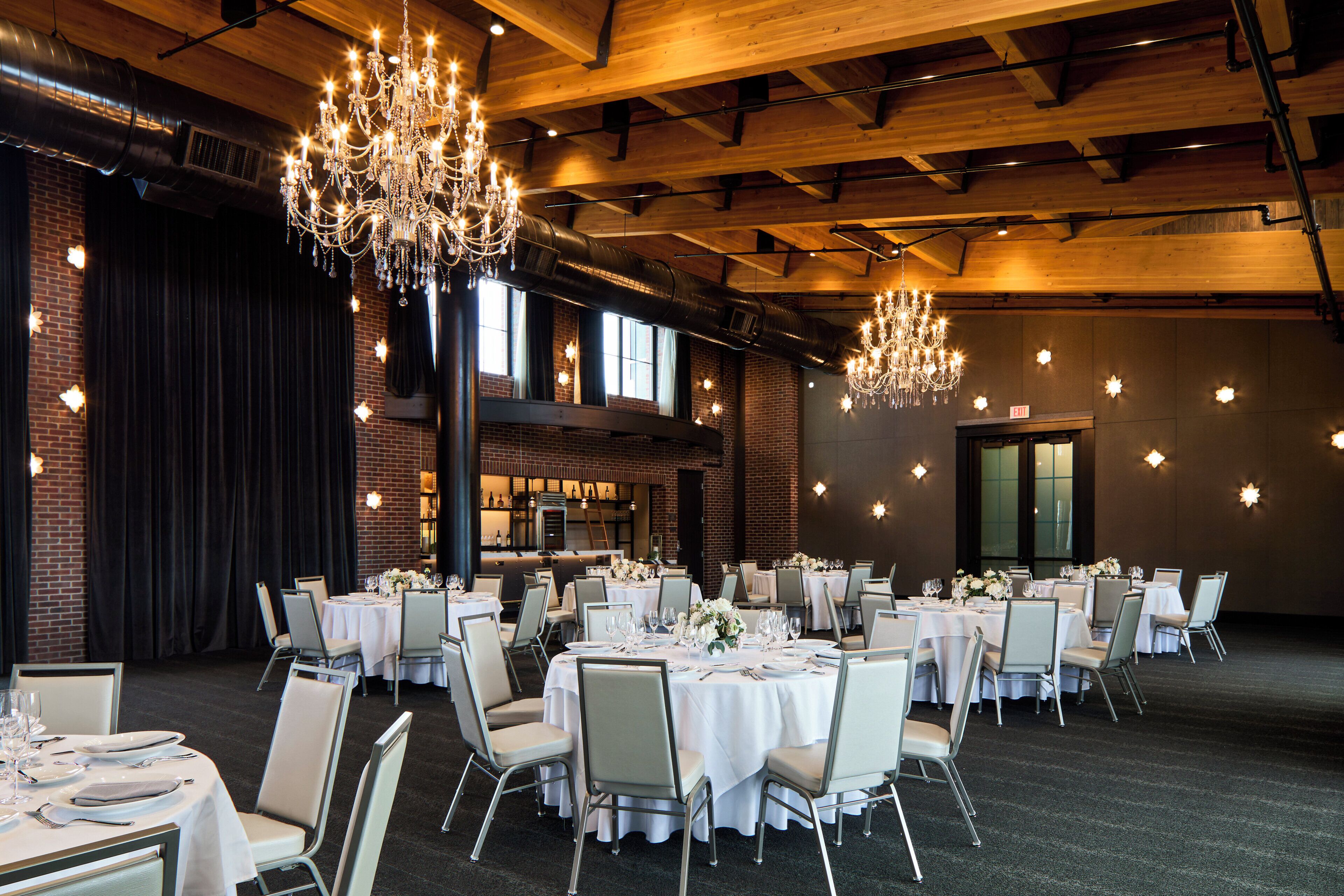 Une salle de banquet avec de nombreuses tables rondes dressées pour le repas, des lustres et des murs en briques.