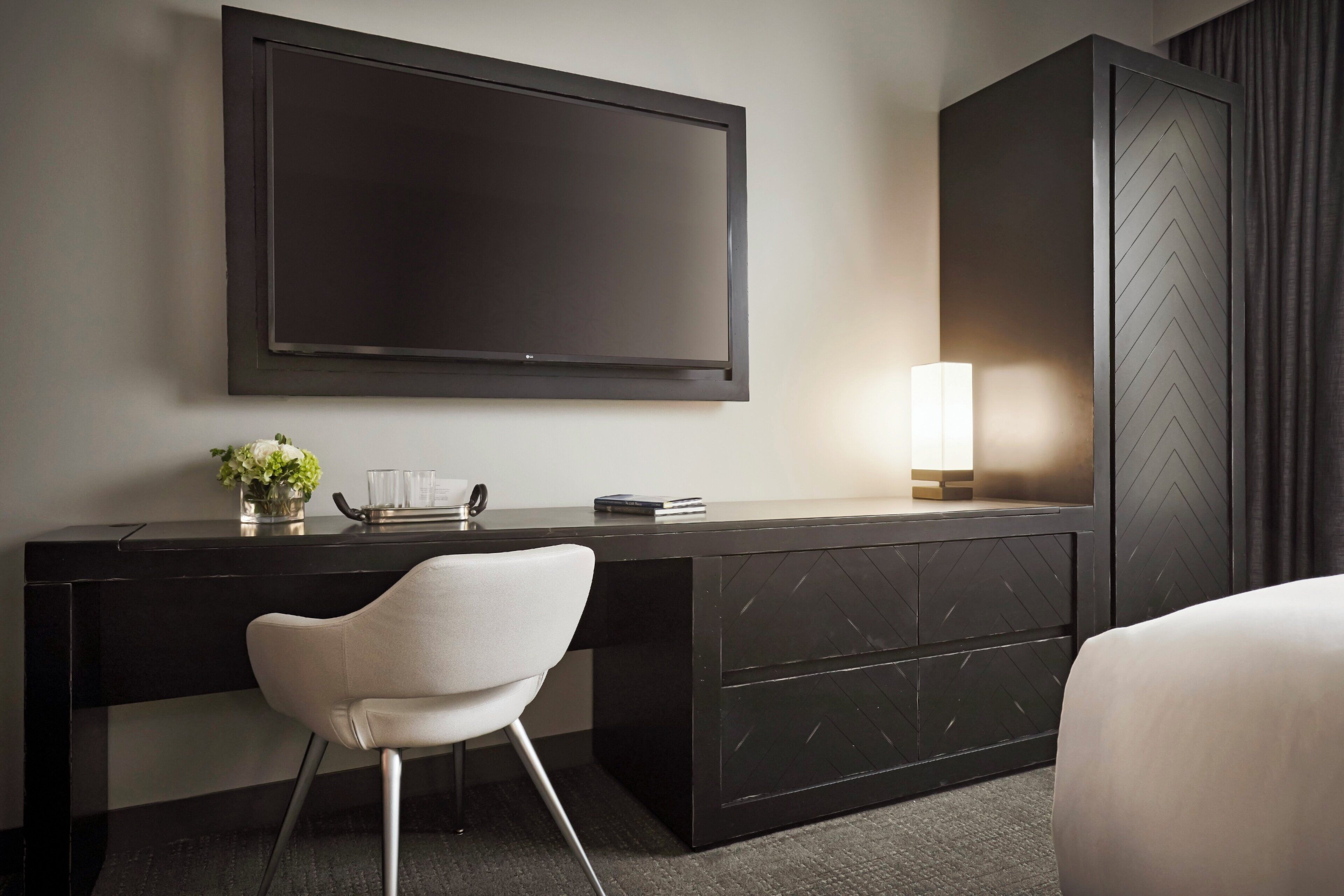 A modern hotel room with a large flat-screen TV mounted on the wall, a desk with a chair, a bedside table with a lamp, and a wardrobe.