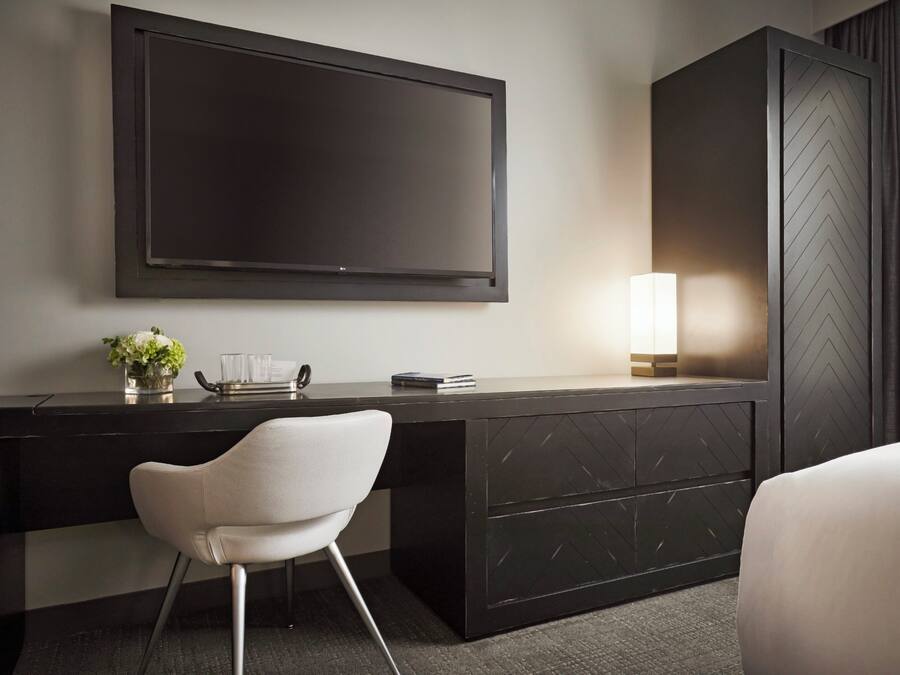 A modern hotel room with a large flat-screen TV mounted on the wall, a desk with a chair, a bedside table with a lamp, and a wardrobe.