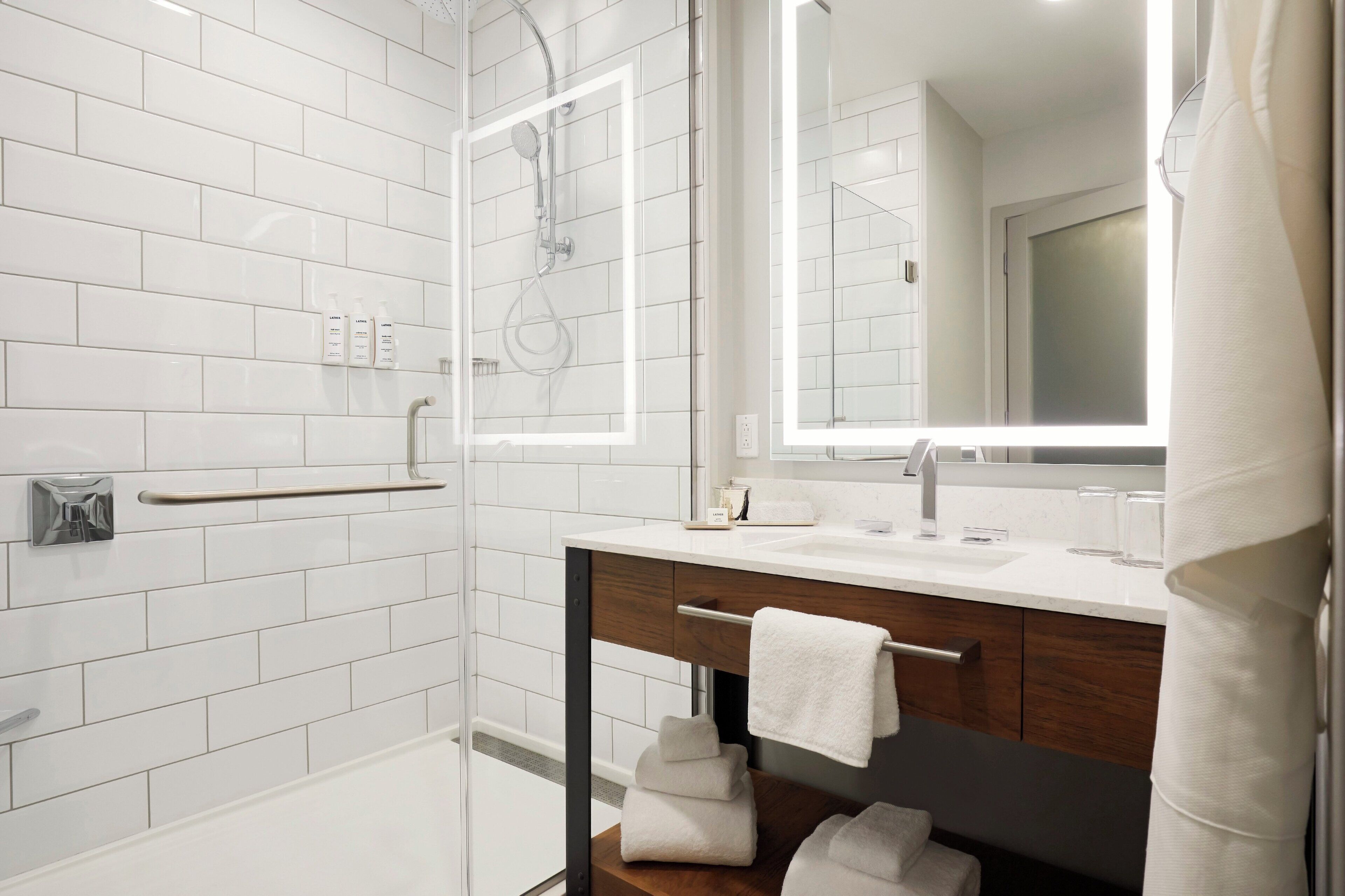 Un baño moderno con azulejos blancos tipo metro, una ducha con cerramiento de vidrio, un mueble de tocador de madera con toallero y un espejo grande.