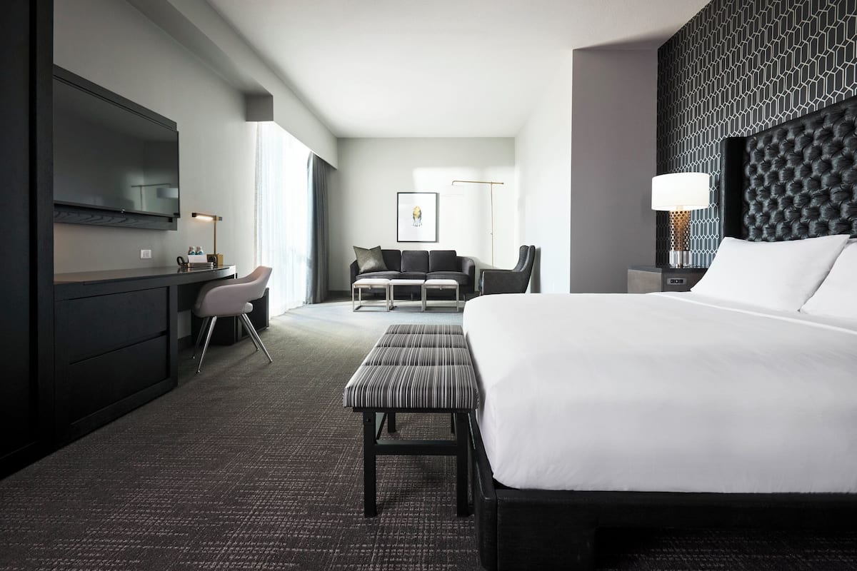 A modern hotel room with a large bed, a desk with a chair, a sofa, and a flat-screen TV mounted on the wall.