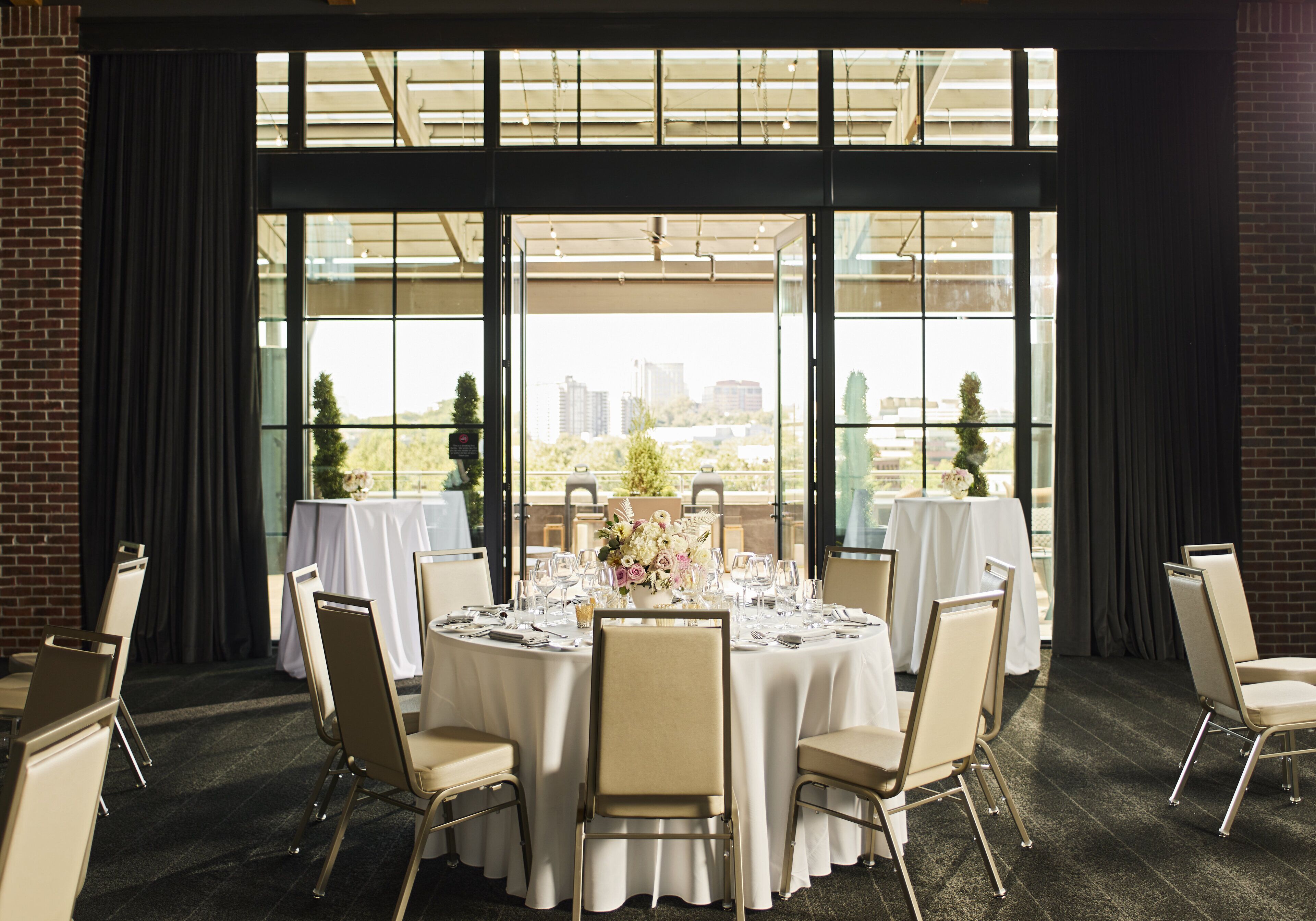 A conference room with a round table set for a meeting, large windows with a city view, and a brick wall.